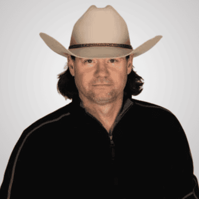 Man wearing a beige cowboy hat and black zip-up jacket, facing the camera on a light gray background.