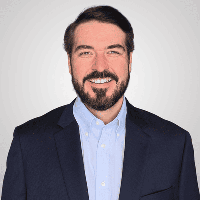 Head-and-shoulders portrait of a smiling man with dark hair and a beard, wearing a navy blazer and light blue shirt.